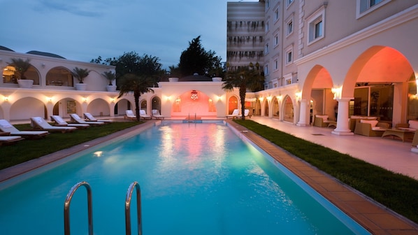 Una piscina con tumbonas y un edificio con pasillos arqueados y mobiliario de exterior.