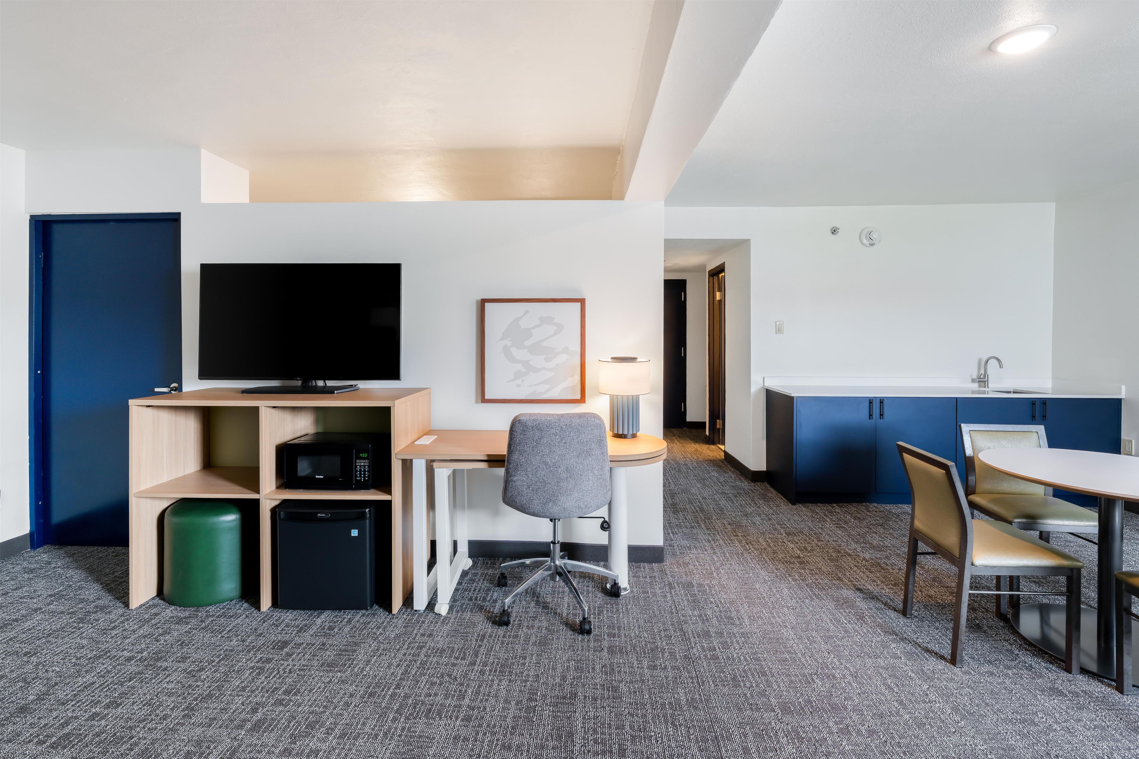 A compact hotel room with a kitchenette, a flat-screen TV, a desk with a chair, and a dining area with a table and chairs.