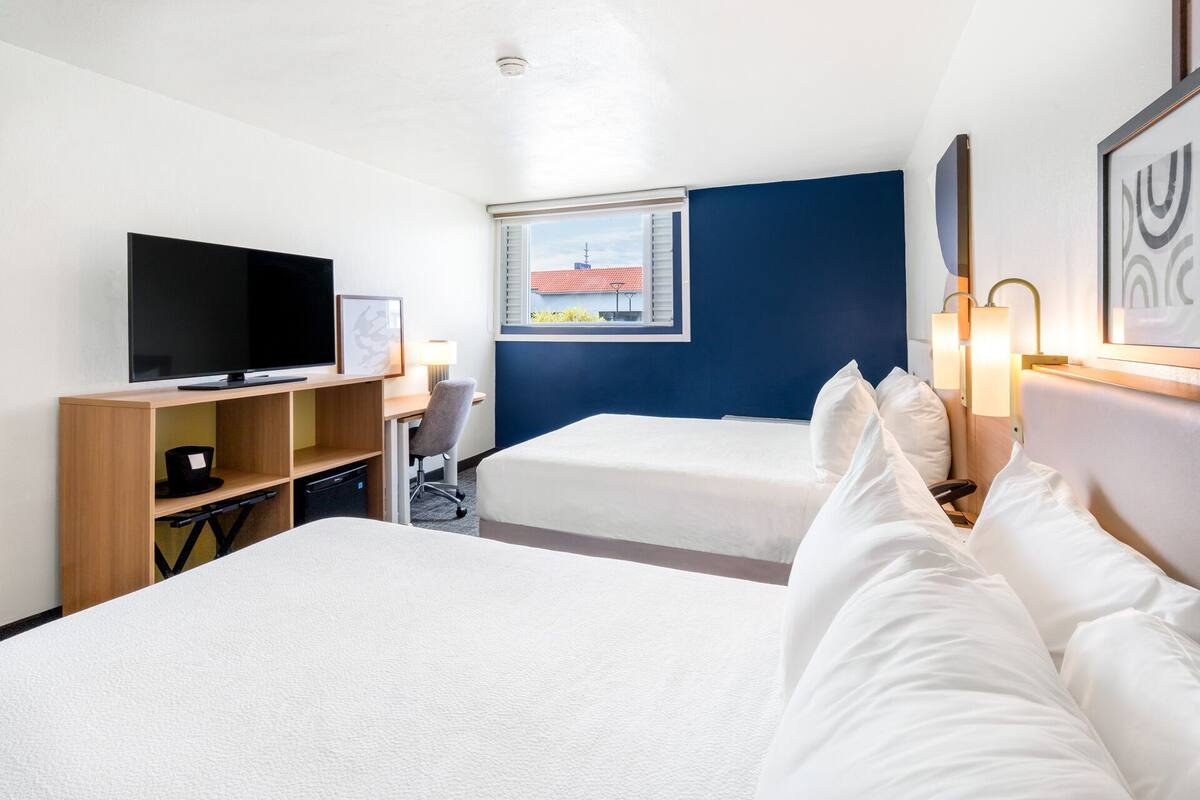 A hotel room with two beds, a TV, a desk, and a window with a view of buildings.