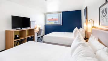 Habitación de hotel con dos camas, televisión, escritorio y ventana con vistas a edificios.