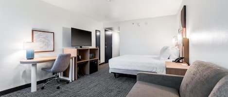 A hotel room with a bed, a desk with a chair, a television, a bookshelf, and a sofa.