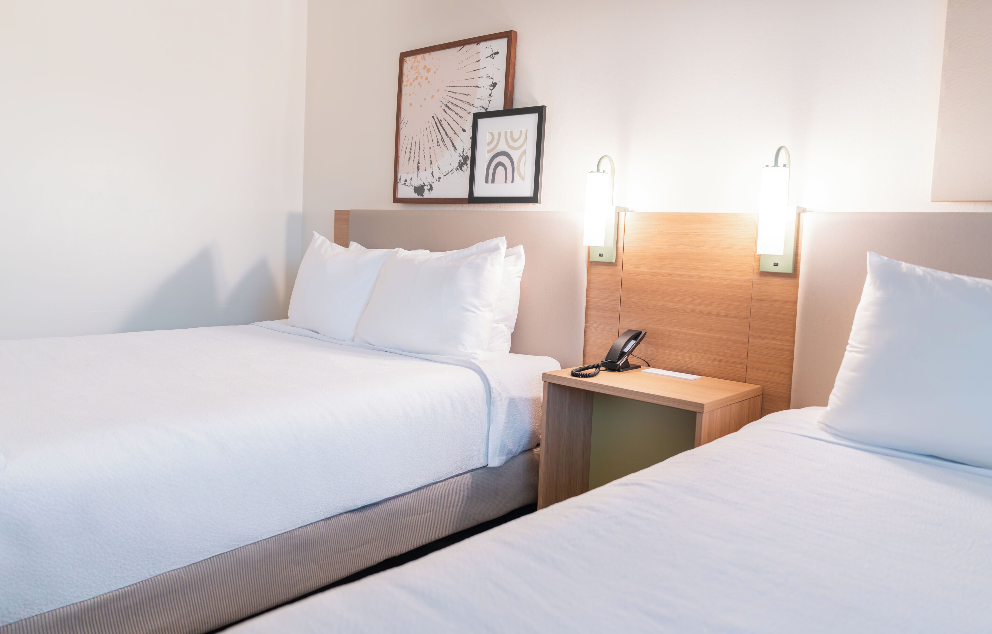 A hotel room with two beds, a bedside table with a phone, and wall-mounted lights.