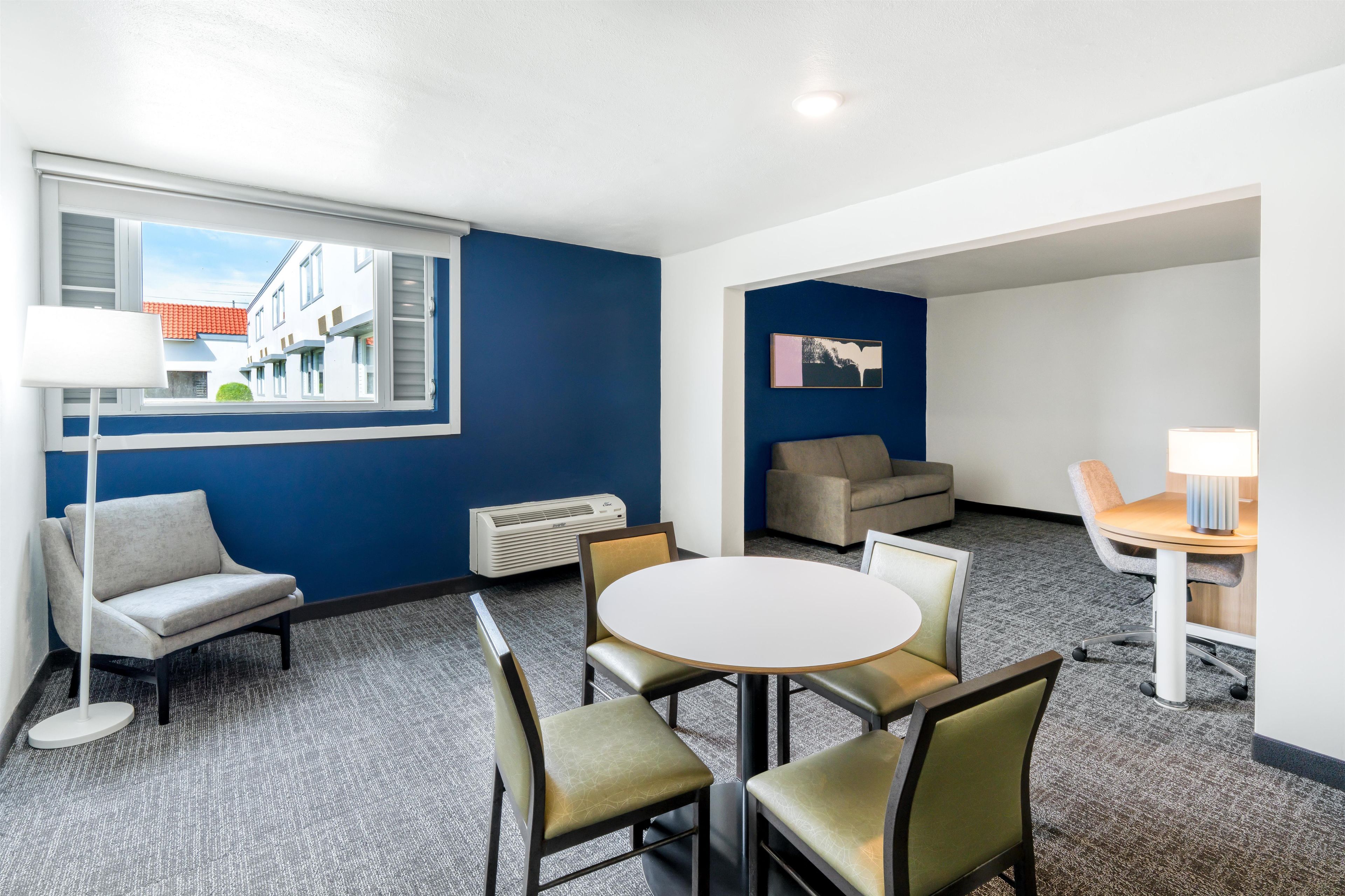 A hotel room with a round table, four chairs, a sofa, a small armchair, a lamp, and a wall-mounted air conditioner.
