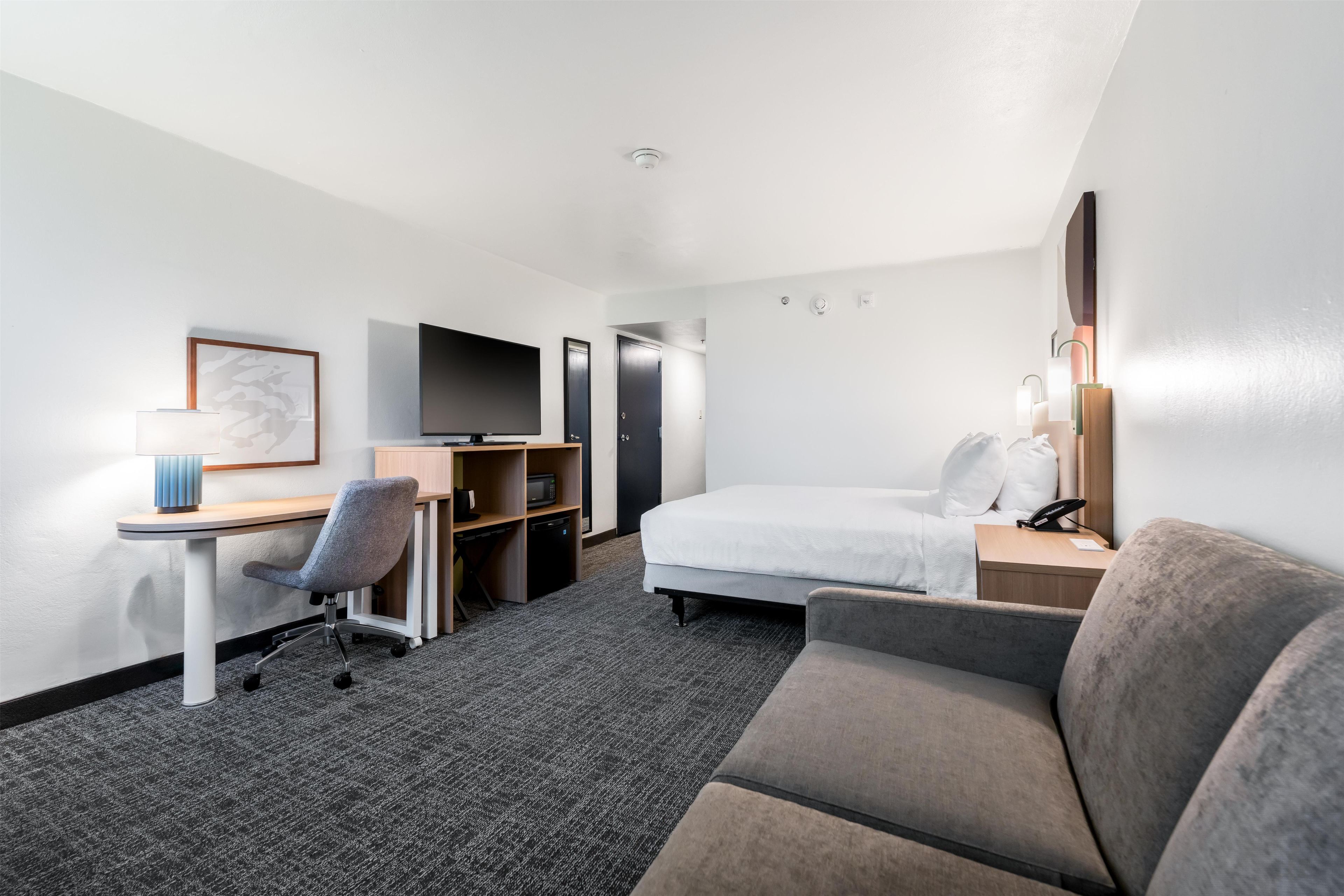 A hotel room with a bed, a desk, a chair, a television, a microwave, and a bedside table with a lamp.