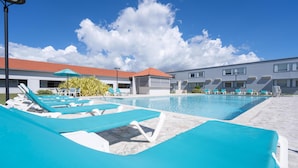 Une piscine entourée de chaises longues bleues, un espace abrité et un bâtiment surmonté d’un toit recouvert de tuiles rouges.
