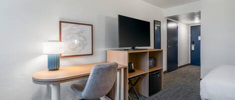 A hotel room with a desk, chair, TV, bed, and a framed picture on the wall.