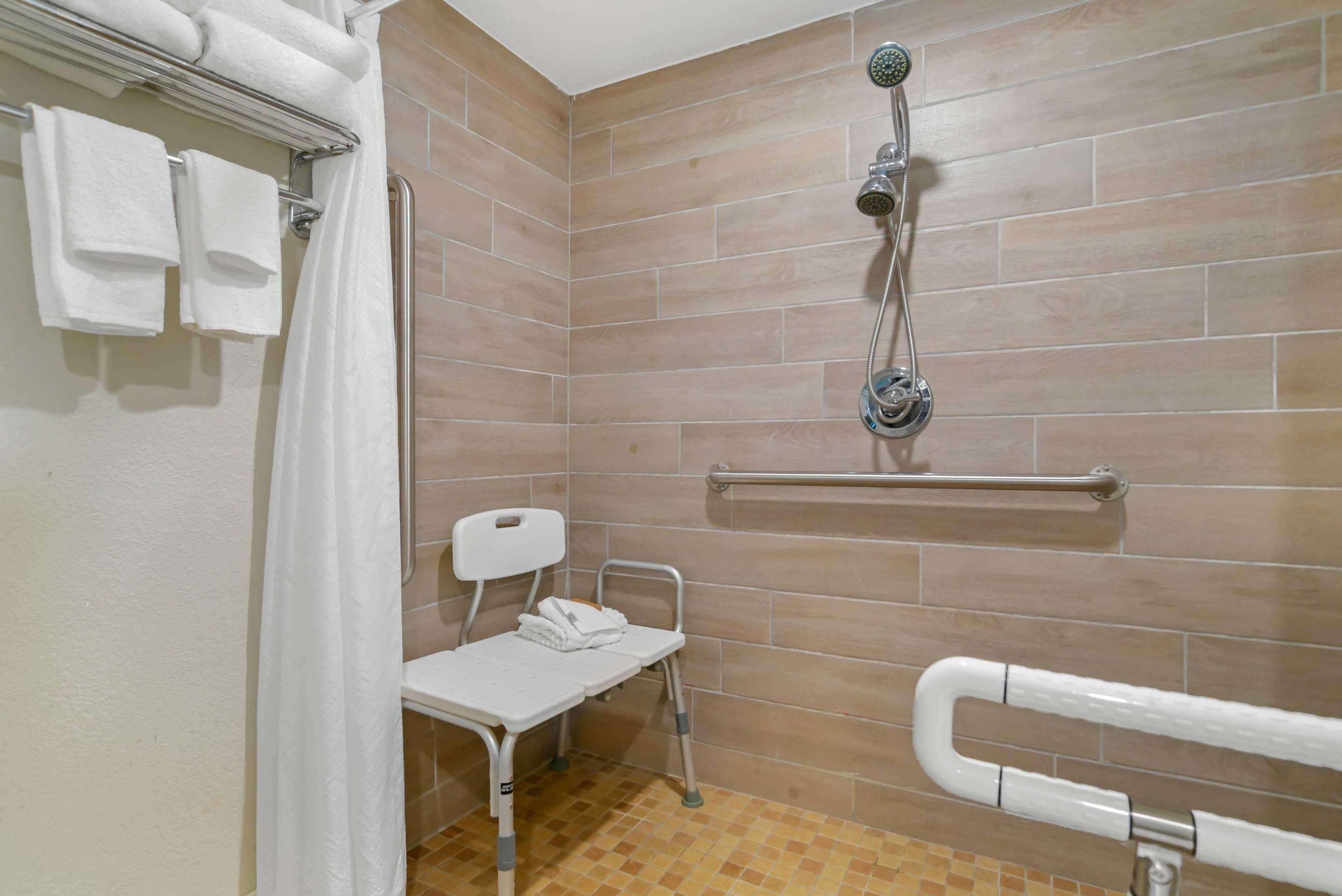 Un baño con ducha, inodoro blanco, barra de apoyo y banco accesible en silla de ruedas.
