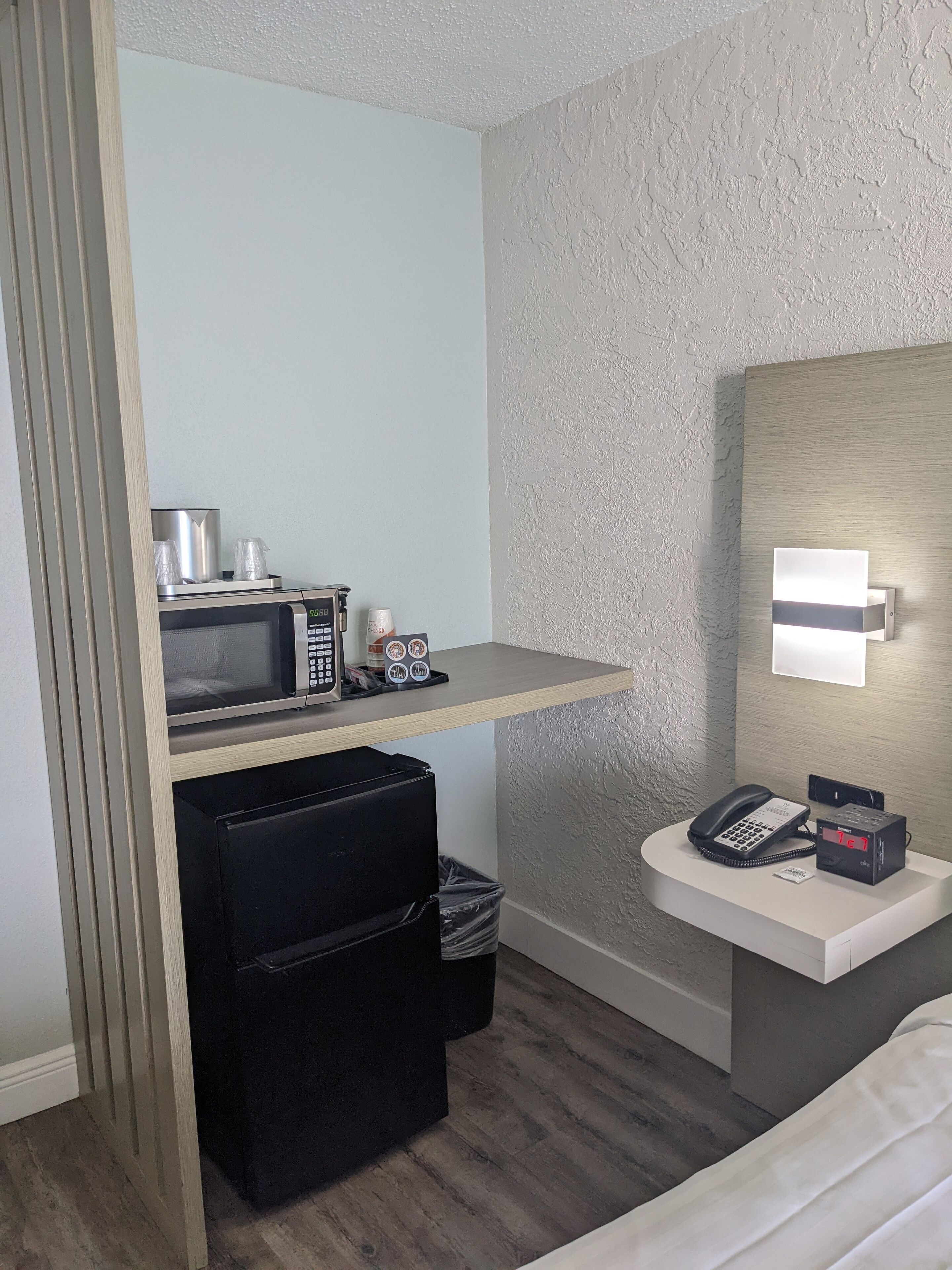 A compact hotel room with a kitchenette, a bedside table with a phone and a digital clock, a small trash bin, and a bed.