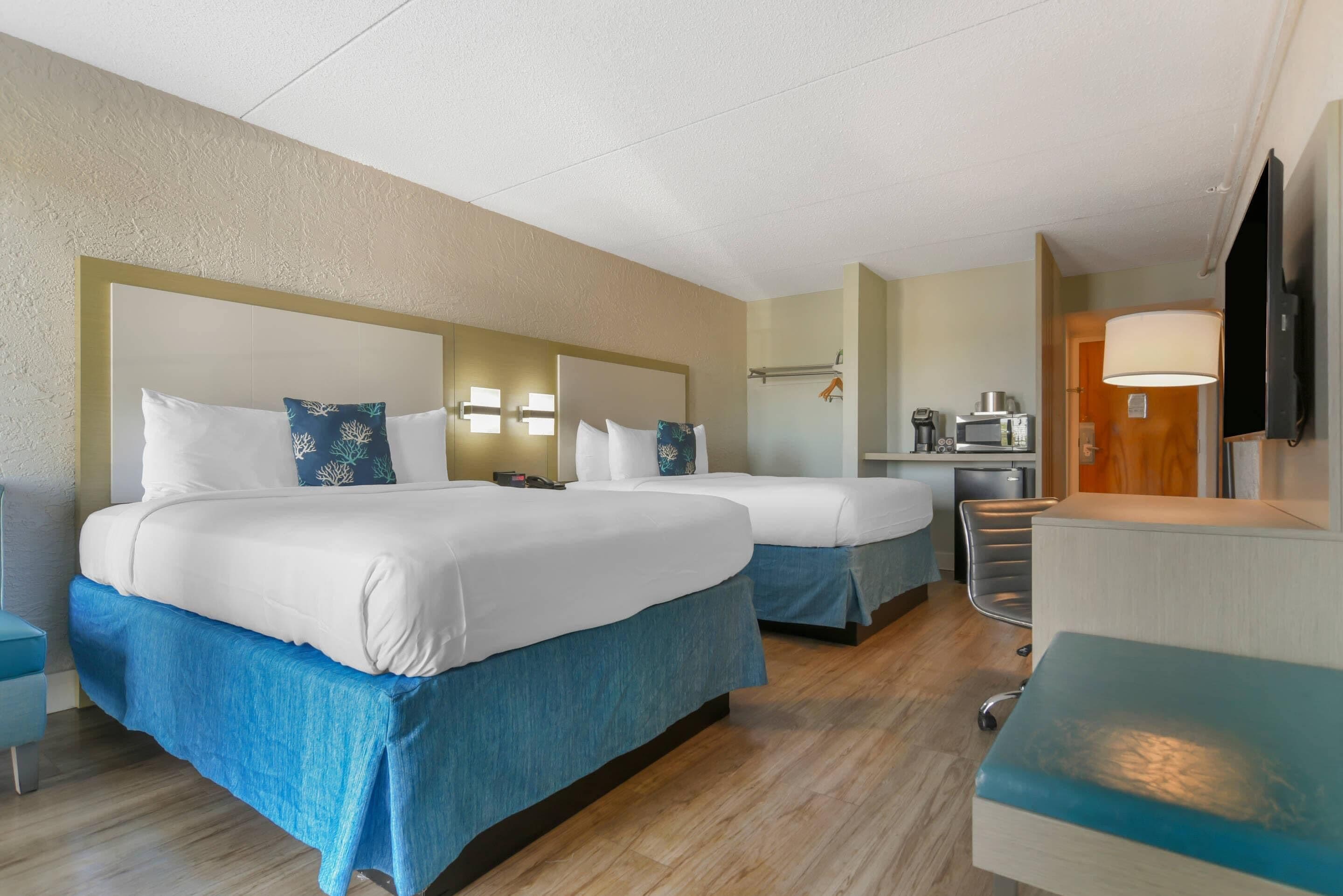 A hotel room with two beds, a desk, a chair, a TV, and a kitchenette.
