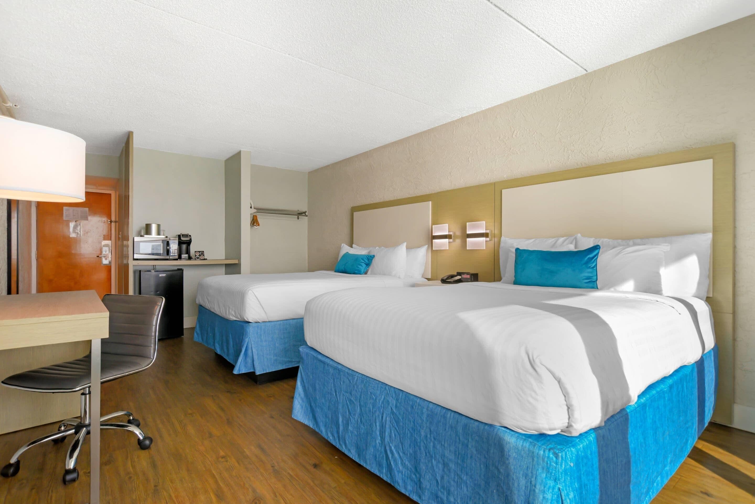 A hotel room with two beds, a desk, a chair, a coffee maker, and a small kitchenette.