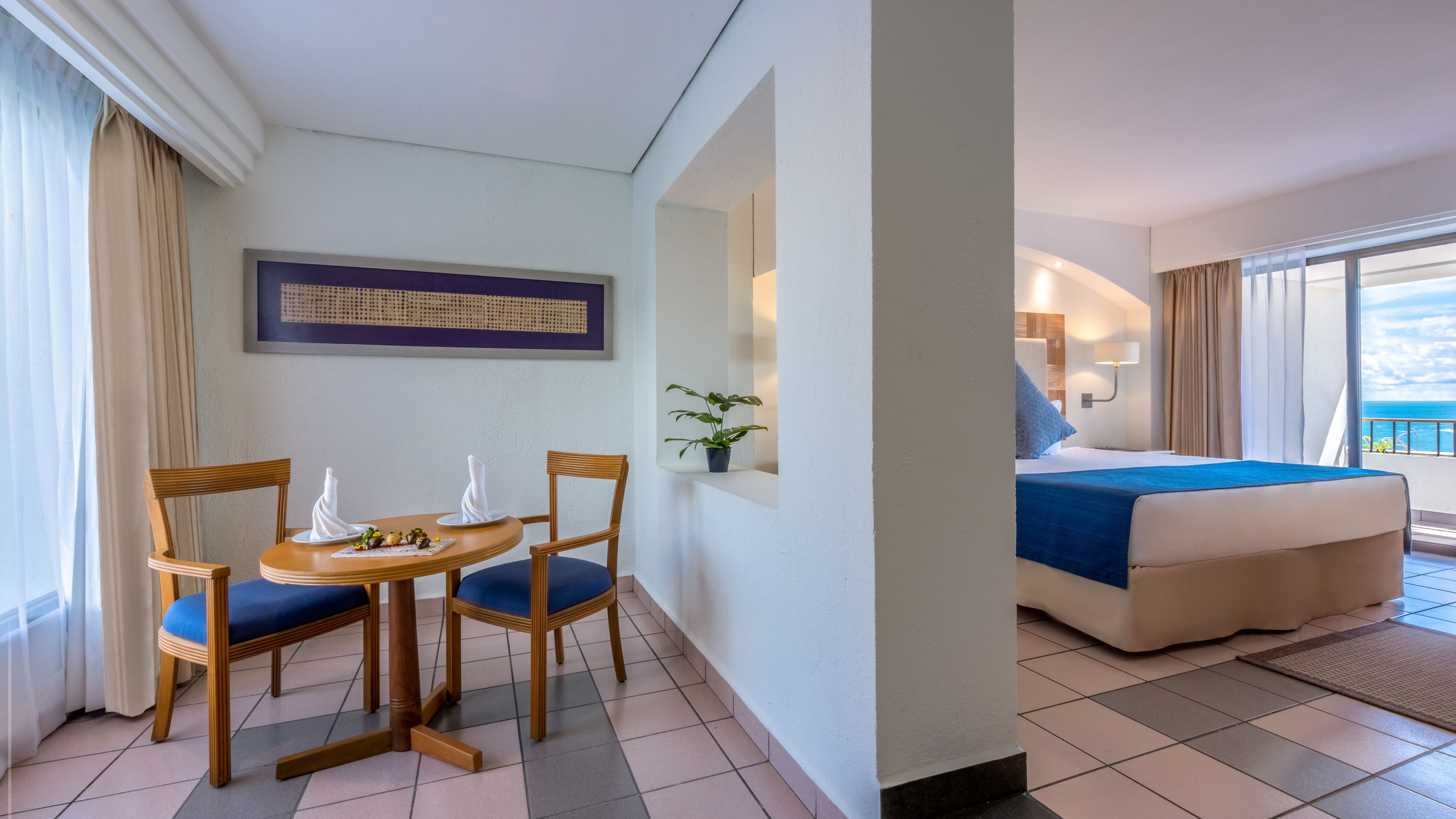 Une chambre d’hôtel avec un lit, une table à manger et des chaises, une plante en pot et une vue sur l’océan.