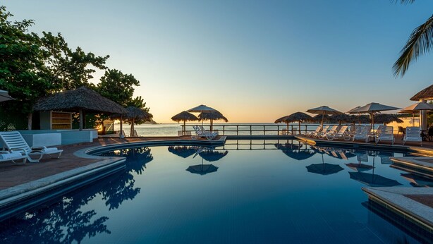 A pool with lounge chairs and umbrellas, a thatched-roof structure, and a view of the sea at sunset.