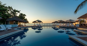 A pool with lounge chairs and umbrellas, a thatched-roof structure, and a view of the sea at sunset.