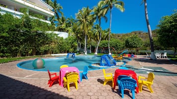 Relax by the serene pool surrounded by lush greenery and palm trees, with comfortable seating and play structures.