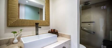 Un baño con un lavamanos grande y rectangular, un espejo con marco texturizado, una planta pequeña y un box de ducha de vidrio.