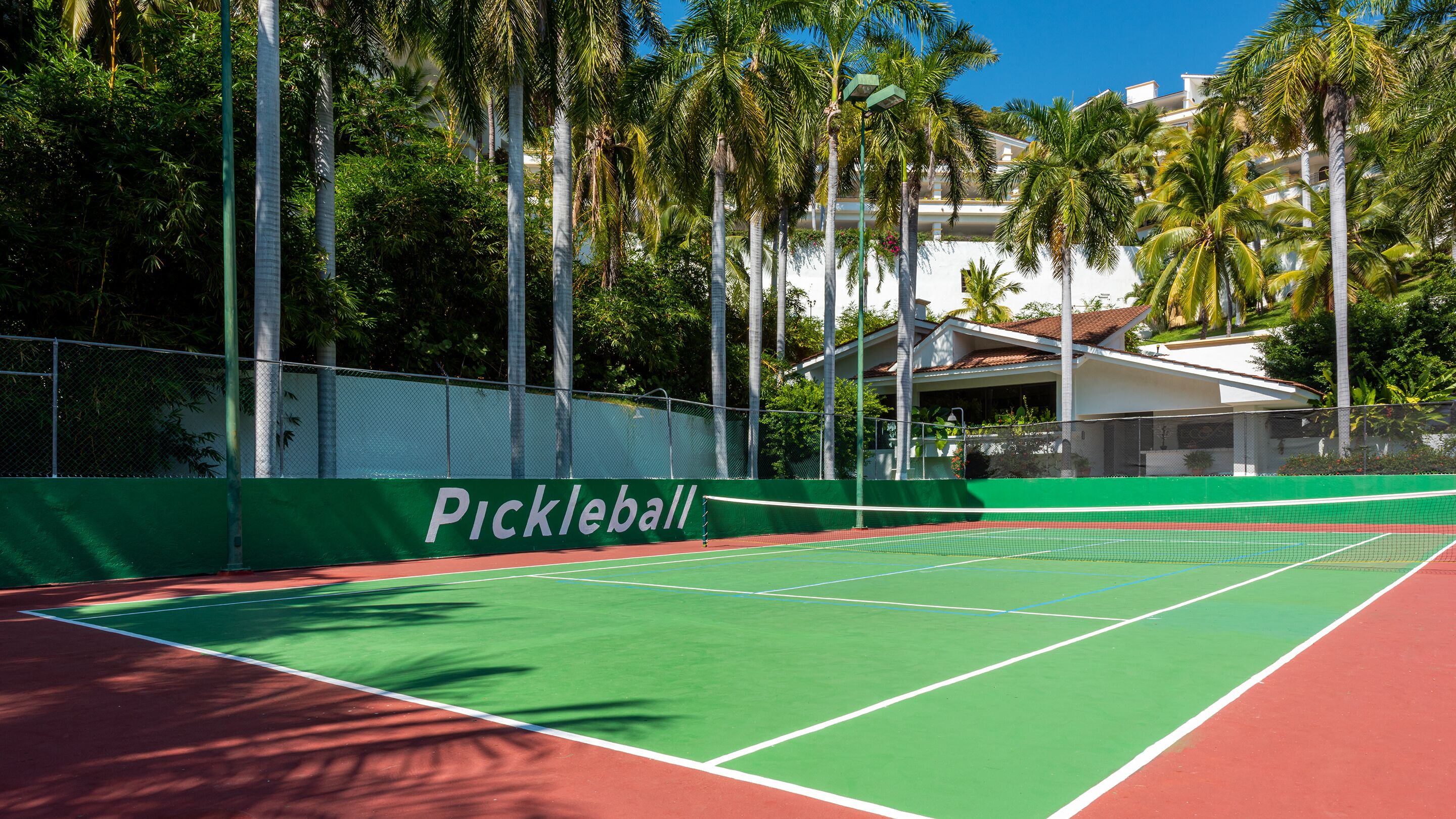 Una cancha de tenis con superficie verde y bordes rojos, rodeada por una valla y palmeras.