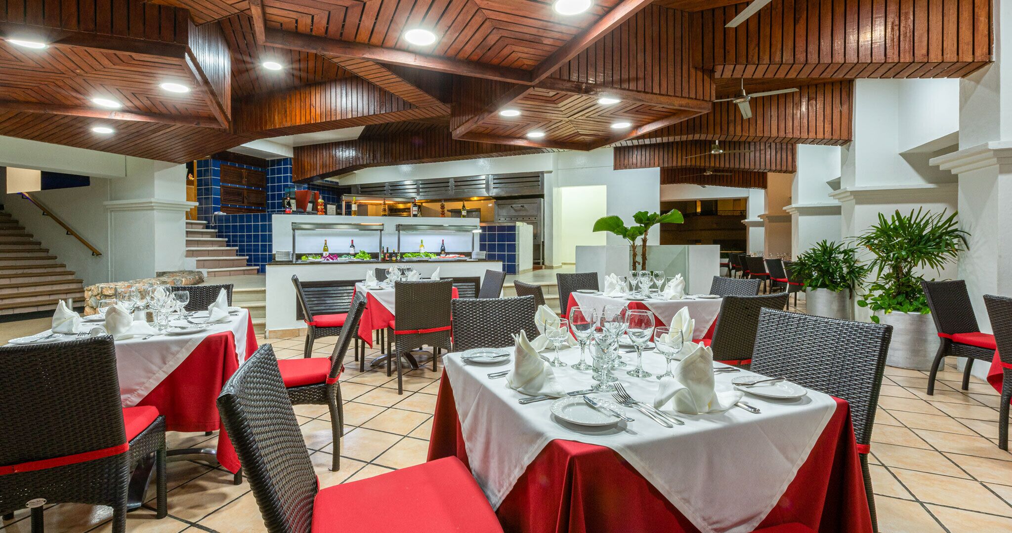 A restaurant with multiple dining tables, wicker chairs, and red cushions. There is a kitchen area in the background with blue tiles and a staircase leading upstairs.