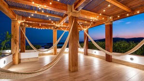 A wooden deck with hammocks and string lights, overlooking a coastal view.
