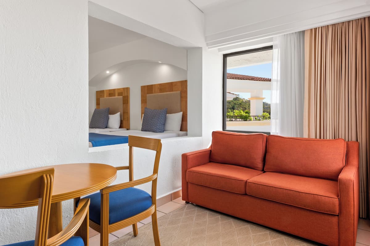Una habitación de hotel moderna con cabecera de madera, sofá rojo, mesa de comedor de madera con cojines azules y vistas al exterior a través de un ventanal grande.