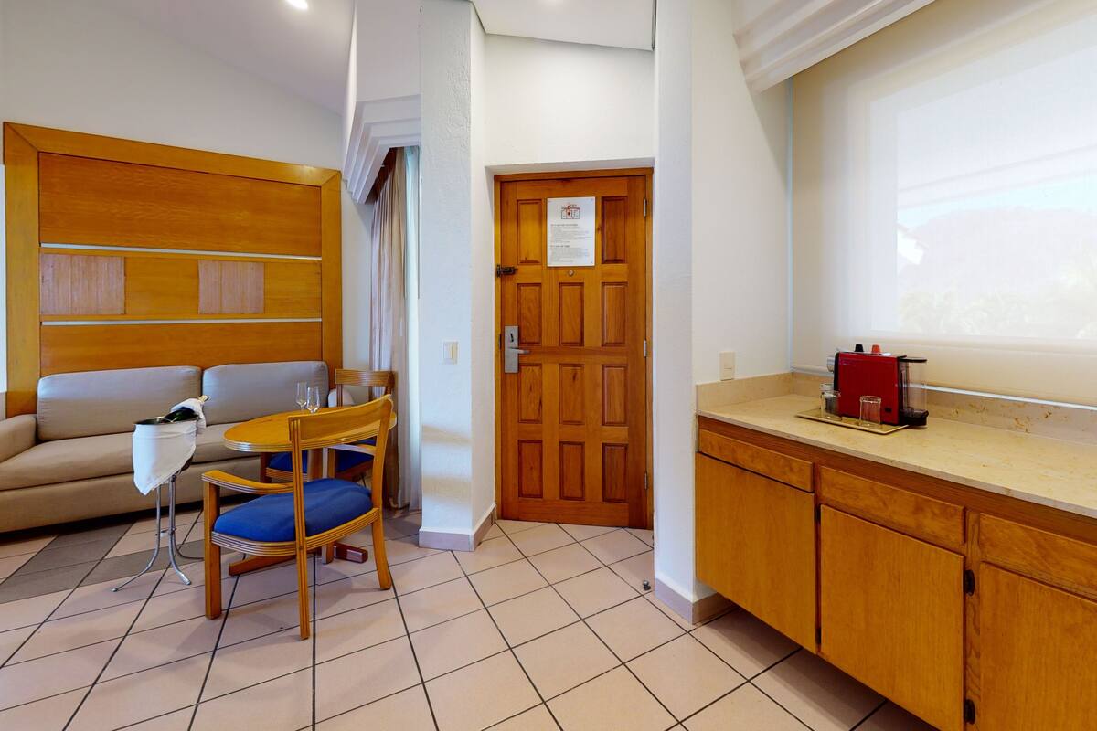 Habitación de hotel con cabecera de madera, un sofá, una mesa de comedor con sillas, una zona de cocina con cafetera y una puerta con letrero.
