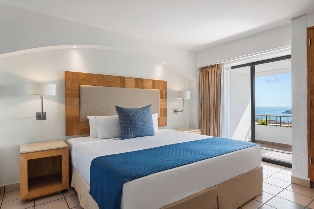 Habitación de hotel con una cama grande, cabecera de madera, dos mesitas de noche con lámparas y vista al océano desde un balcón.