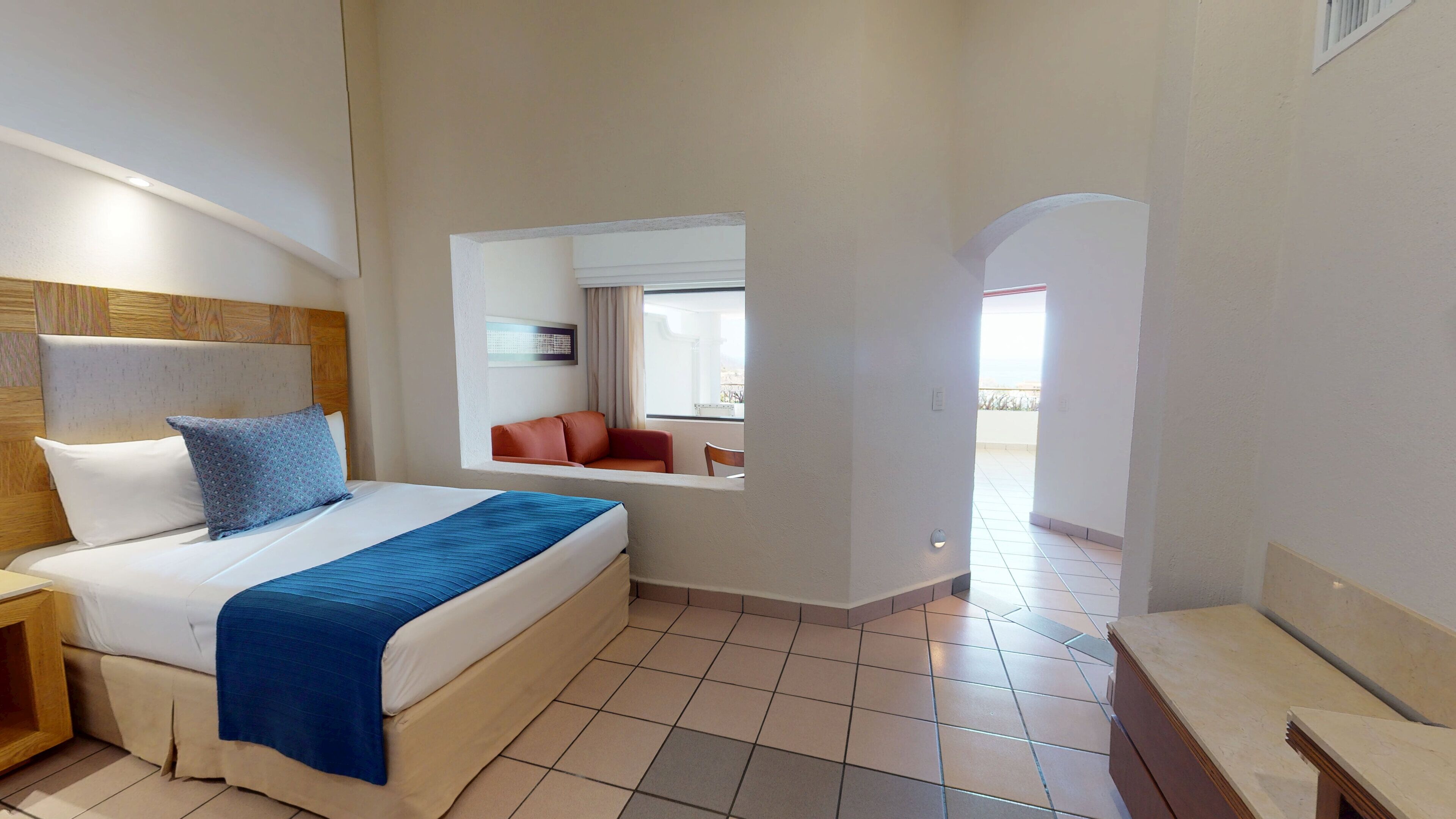 Habitación de hotel con cama, mesita de noche, una silla roja y vistas al balcón.