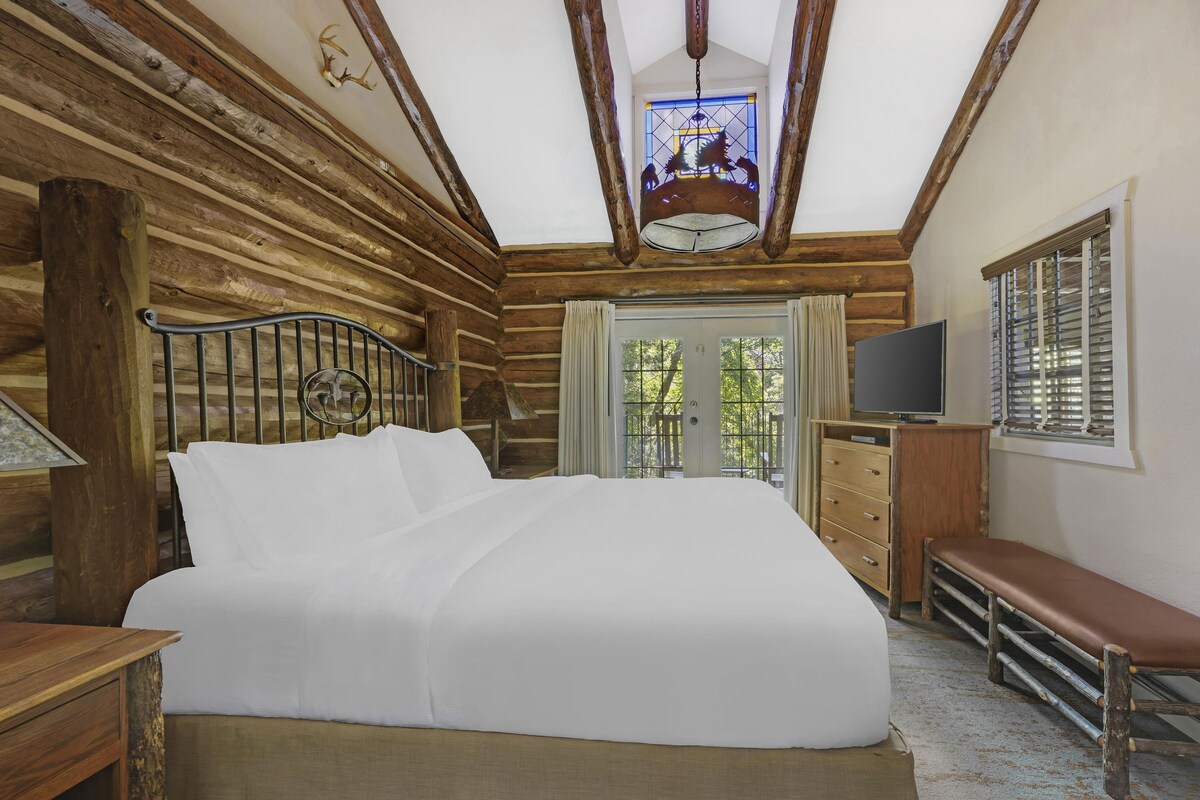 A log cabin bedroom with a large bed, a wooden bench, a television, and a window with blinds.