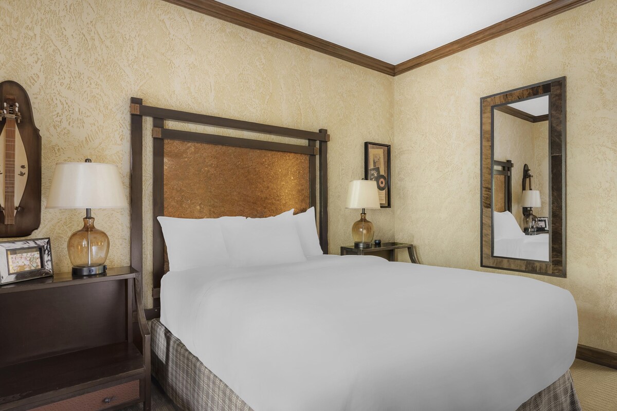 A hotel room with a large bed, two bedside tables with lamps, a wooden headboard, a wall-mounted mirror, and a framed picture.