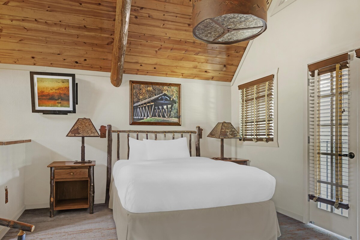 A bedroom with a wooden ceiling, a bed, two nightstands, two lamps, a framed picture of a covered bridge, and a window with blinds.
