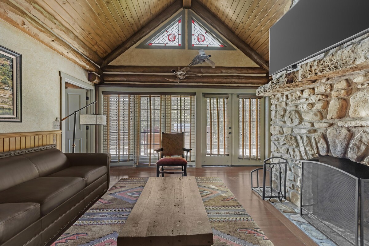 A spacious room with a wooden ceiling, a stone wall, a fireplace, a sofa, and a patterned rug.