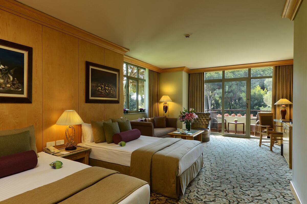 Quarto de hotel com duas camas, um sofá, área de refeições com mesa e cadeiras, e janelas amplas com vista para o exterior.