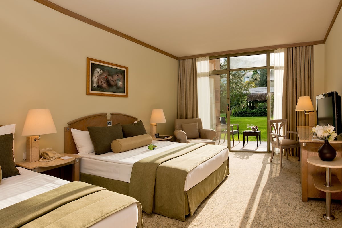Quarto de hotel com duas camas, uma mesa de trabalho com televisão, uma cadeira e vista para o jardim.