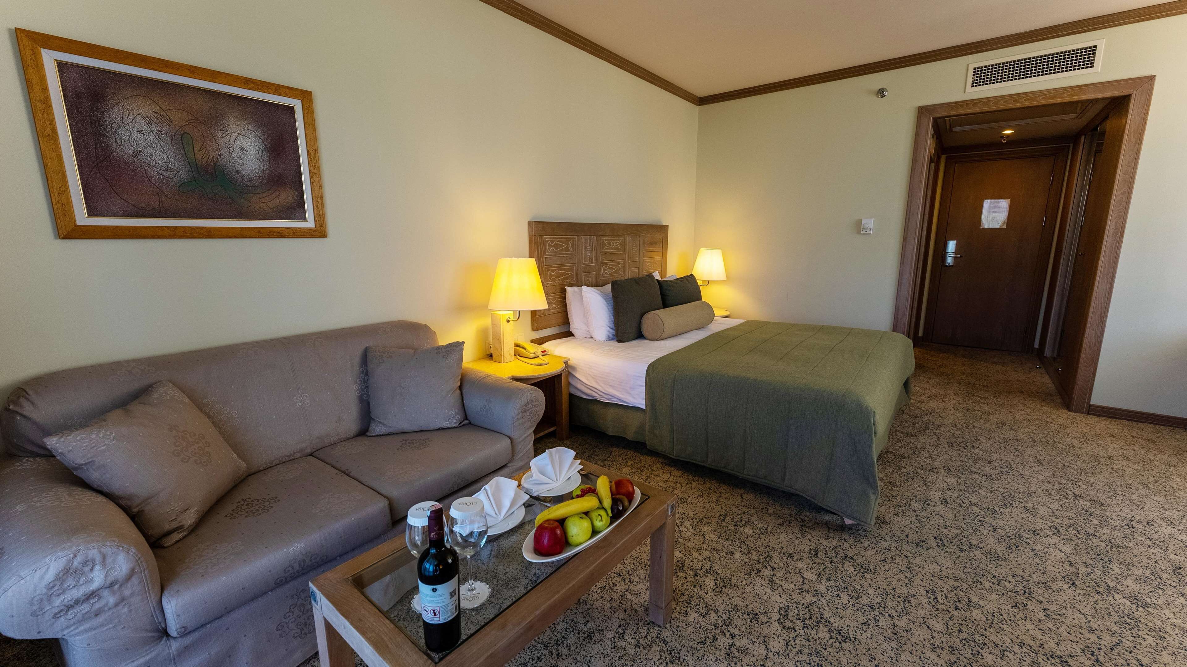 Hotelkamer met een bank, een bed met een groene plaid, een nachtkastje met een lamp, een klein tafeltje met een fles wijn en fruit, en een schilderij aan de muur.