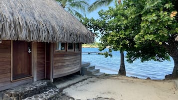 A wooden building with a thatched roof, a stone staircase leading to a sandy beach, and a palm tree.