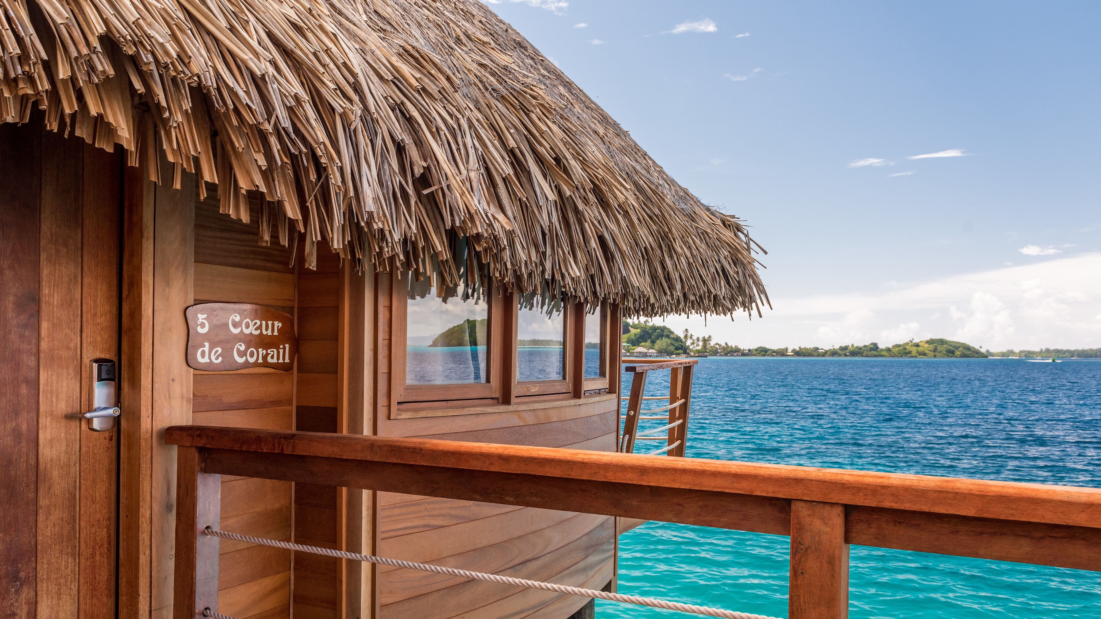 Una cabaña de madera sobre el agua con techo de paja, una terraza de madera y un letrero que dice "5 Coeur de Corail".