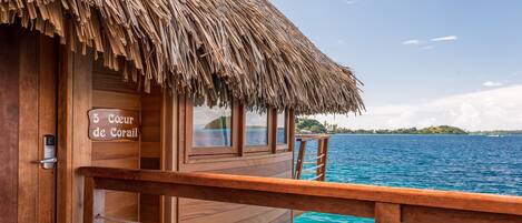 A wooden overwater bungalow with a thatched roof, a wooden deck, and a sign reading "5 Coeur de Corail".