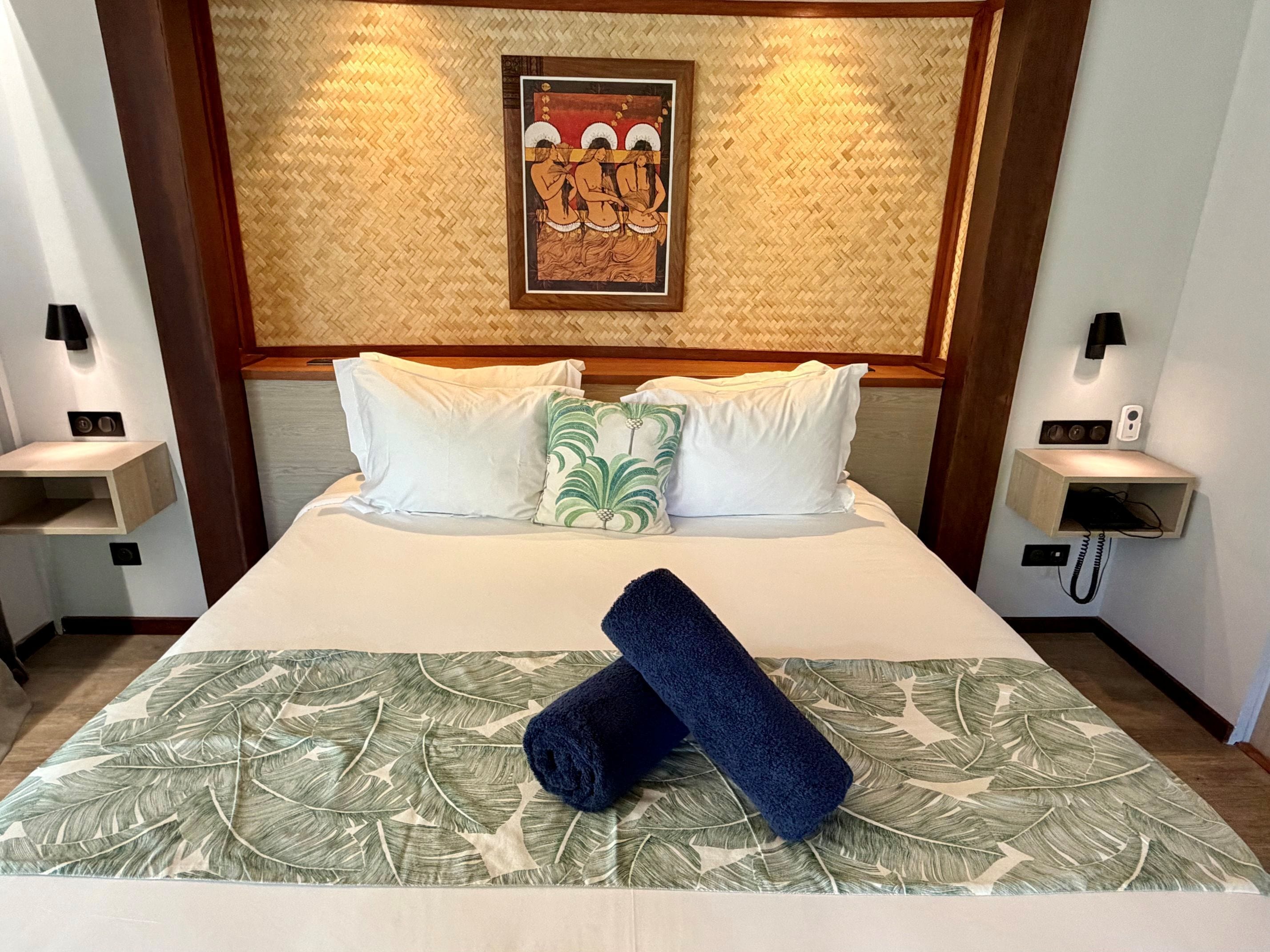 Habitación de hotel con una cama grande, ropa de cama blanca, colcha de hojas verdes, toalla azul y una obra de arte enmarcada sobre la cama.