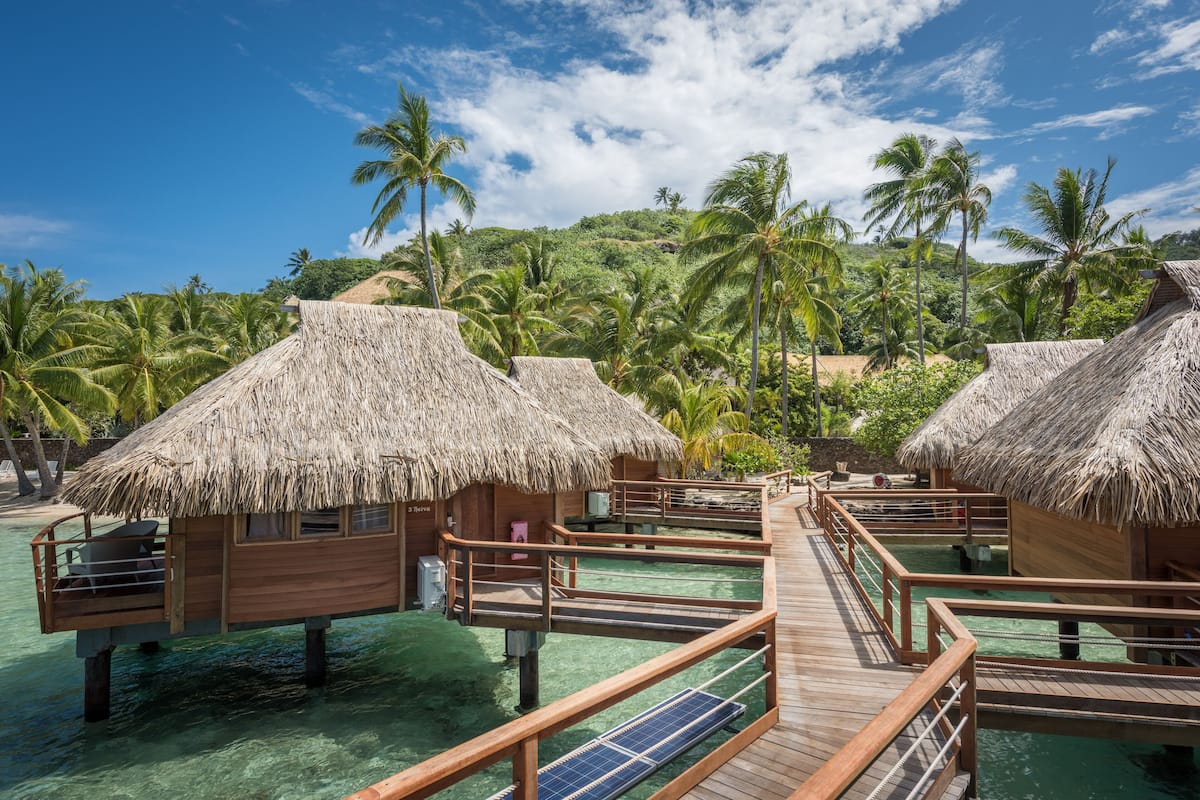 Overwater bungalows with thatched roofs, wooden walkways, and clear turquoise water.