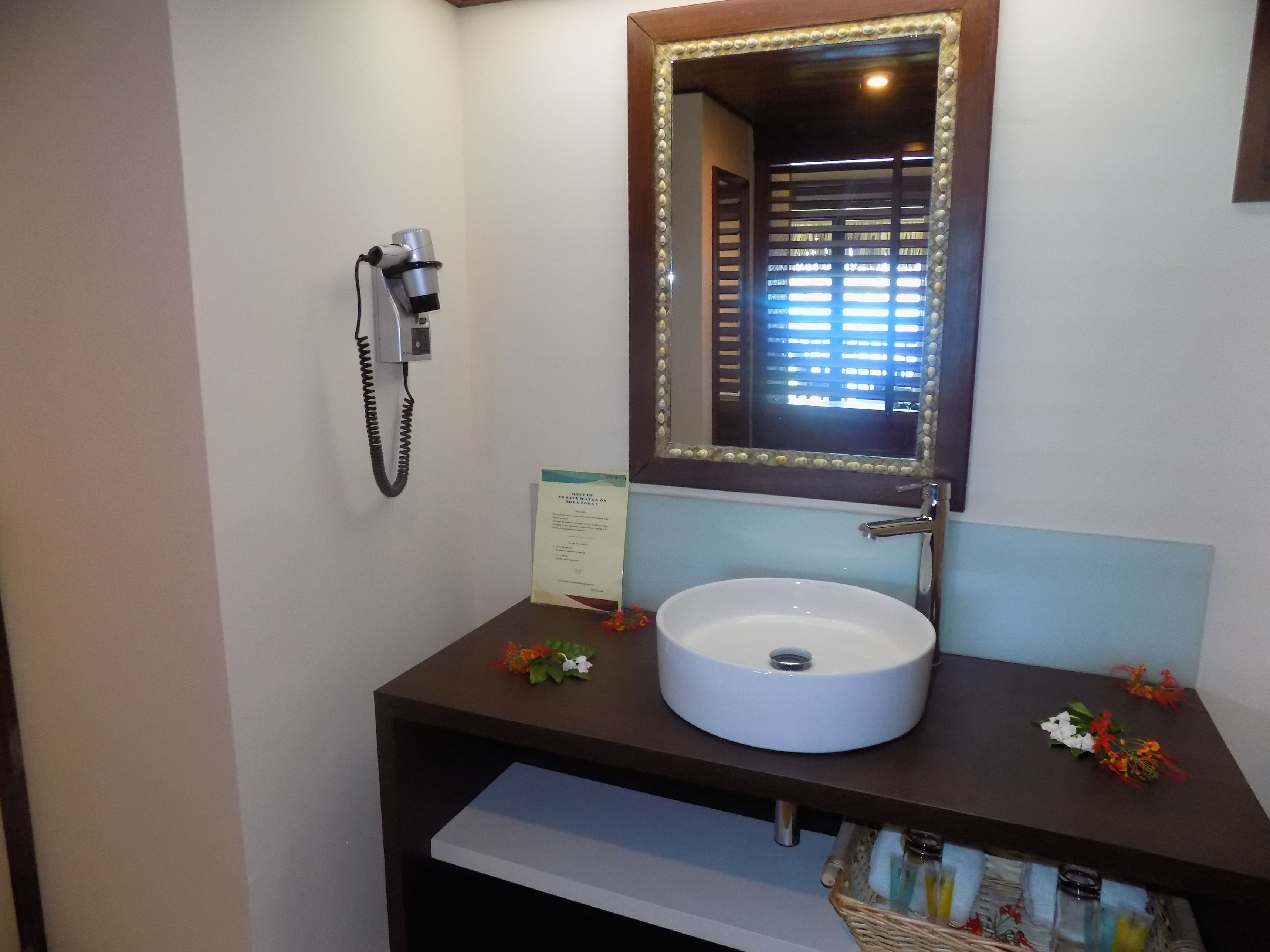 Een badkamer met een witte wastafel, een spiegel met decoratieve lijst, een haardroger en een mandje met toiletartikelen.