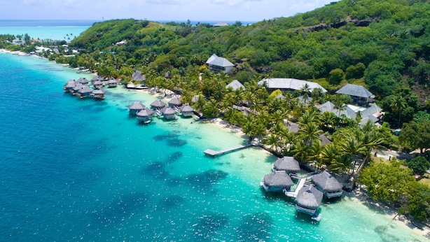 Vista aérea de un resort tropical con bungalows sobre el agua, aguas turquesas cristalinas y abundante vegetación.