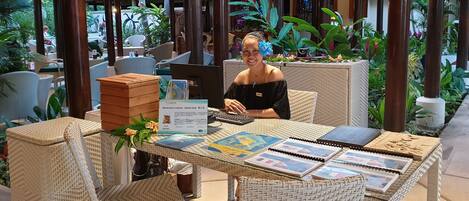 Una mujer sentada a una mesa con una computadora, rodeada de muebles de mimbre y plantas en un espacio al aire libre.