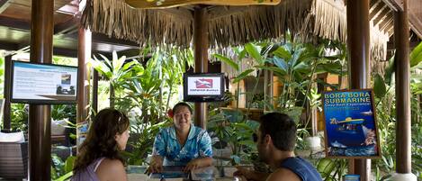 A reception desk with a wooden structure, tropical plants, and informational signs about hotel activities.