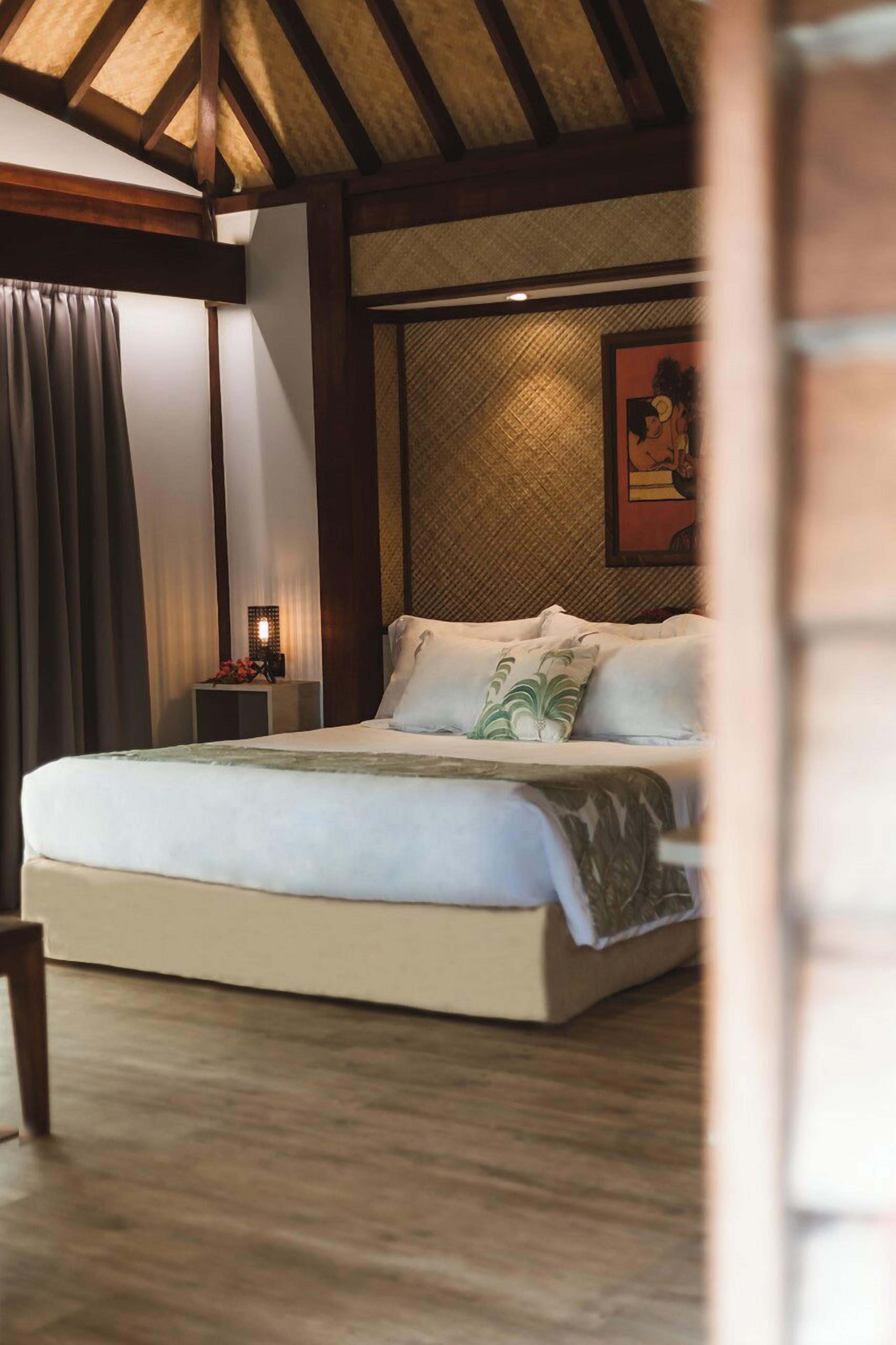 A bedroom with a large bed, wooden beams on the ceiling, a nightstand with a lamp, and a framed picture on the wall.