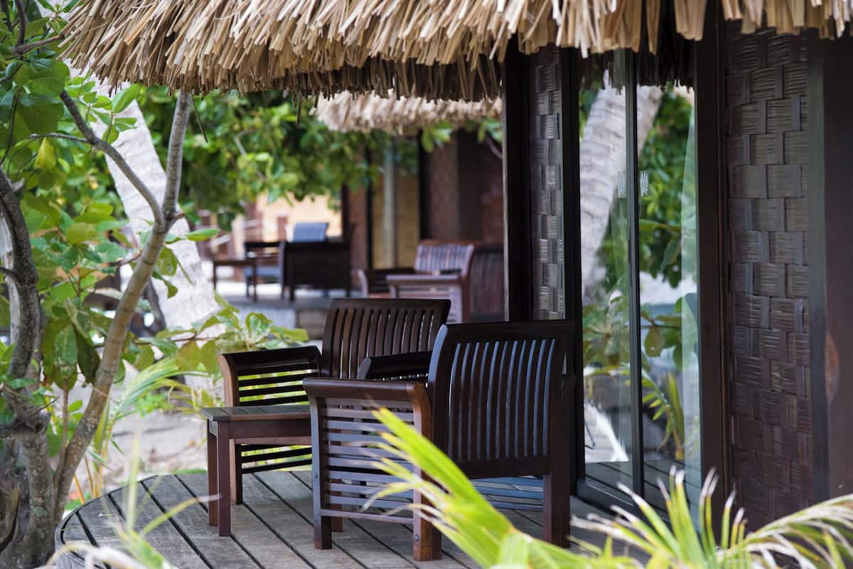 A wooden deck with chairs and a thatched roof structure, surrounded by greenery.