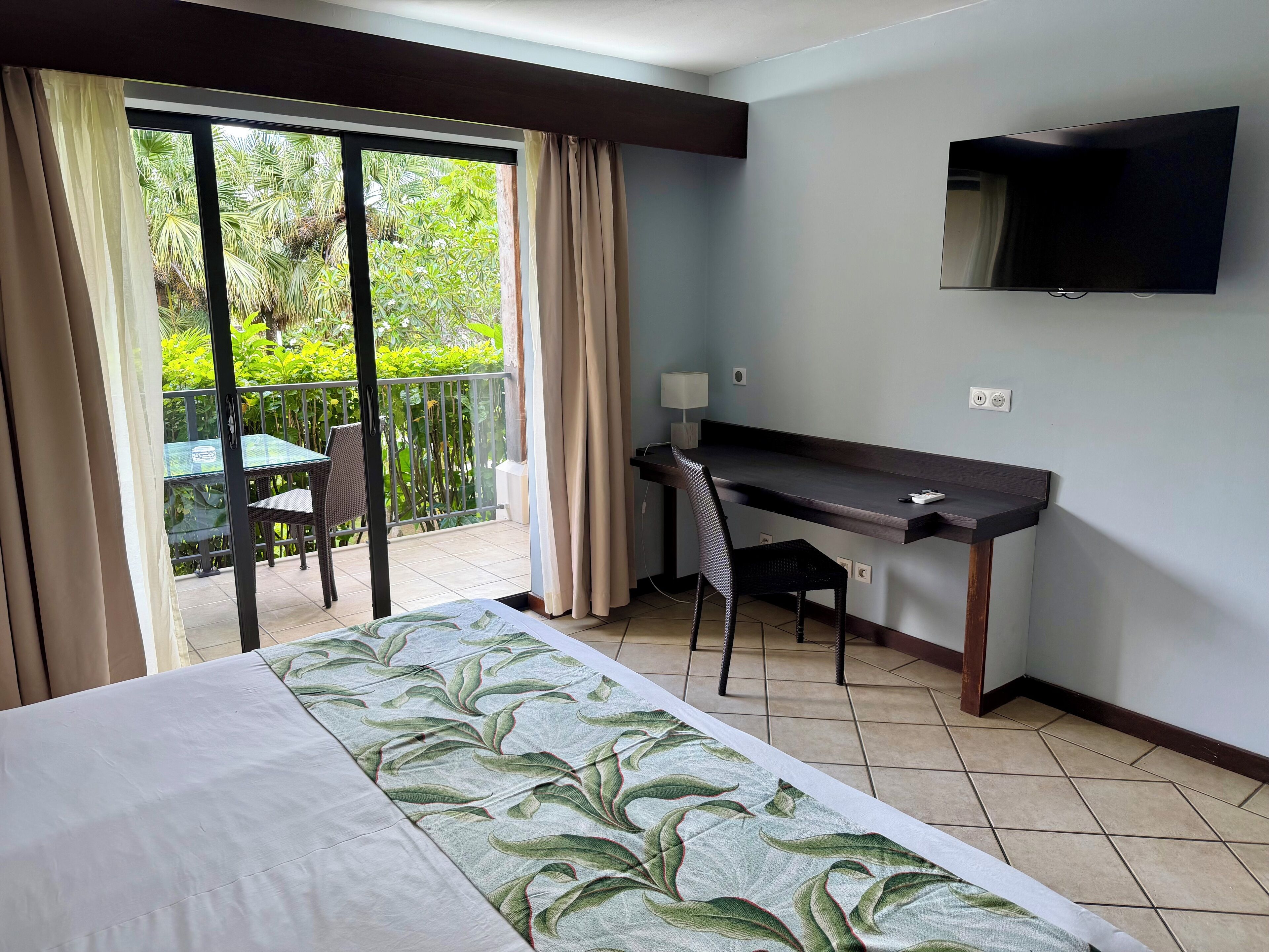 Habitación de hotel con cama, escritorio, silla, televisión y balcón con mesa y sillas.