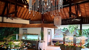 Amplio espacio interior con techo alto, vigas de madera y un candelabro decorativo. Se observan estaciones tipo buffet con decoración floral y un fondo de pared de piedra.