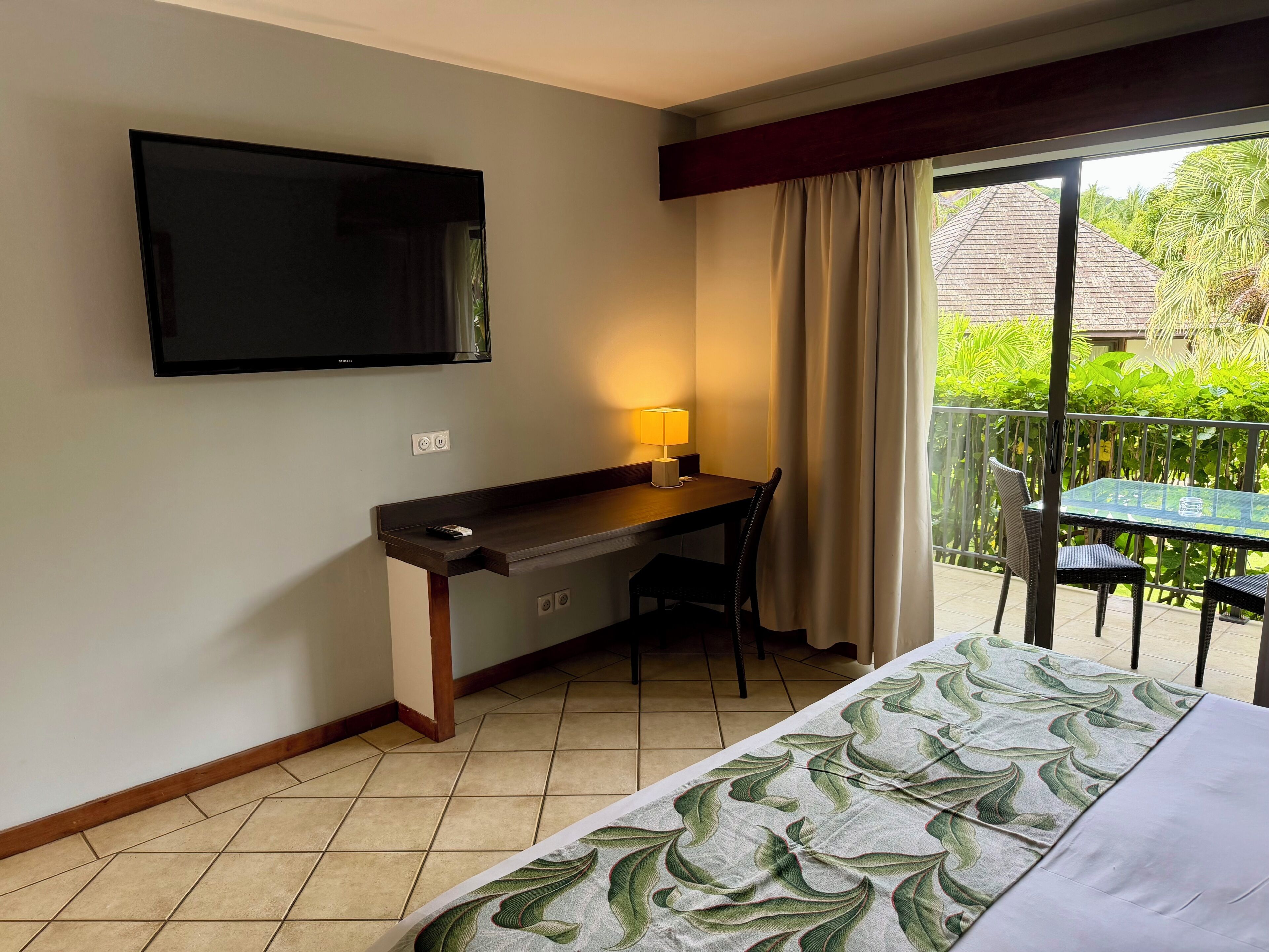 Habitación de hotel con cama, escritorio con silla, televisor, balcón con mesa y sillas, y vistas a zonas verdes.
