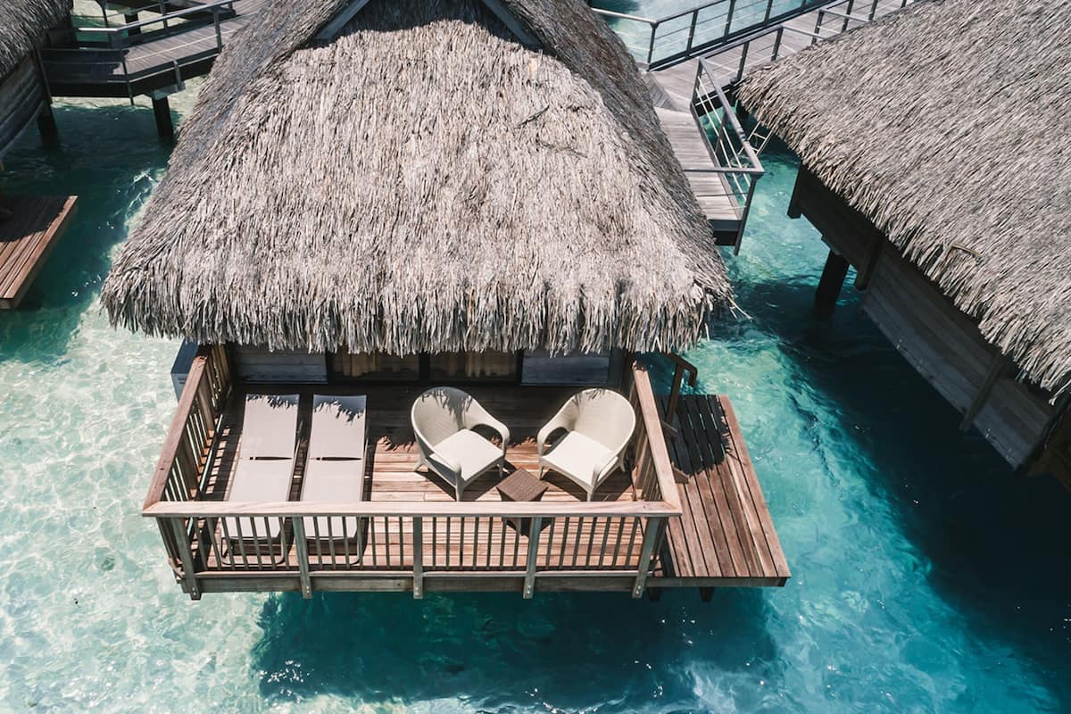 An overwater bungalow with thatched roofs, wooden deck, and two lounge chairs.