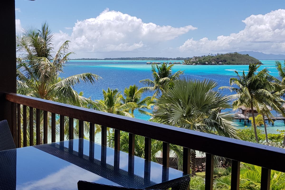 A balcony with a glass table and chairs, overlooking a turquoise sea and a tropical island.
