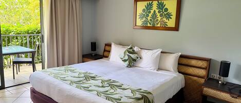 A hotel room with a bed, a nightstand, a lamp, a table with chairs, and a view of greenery through a window.